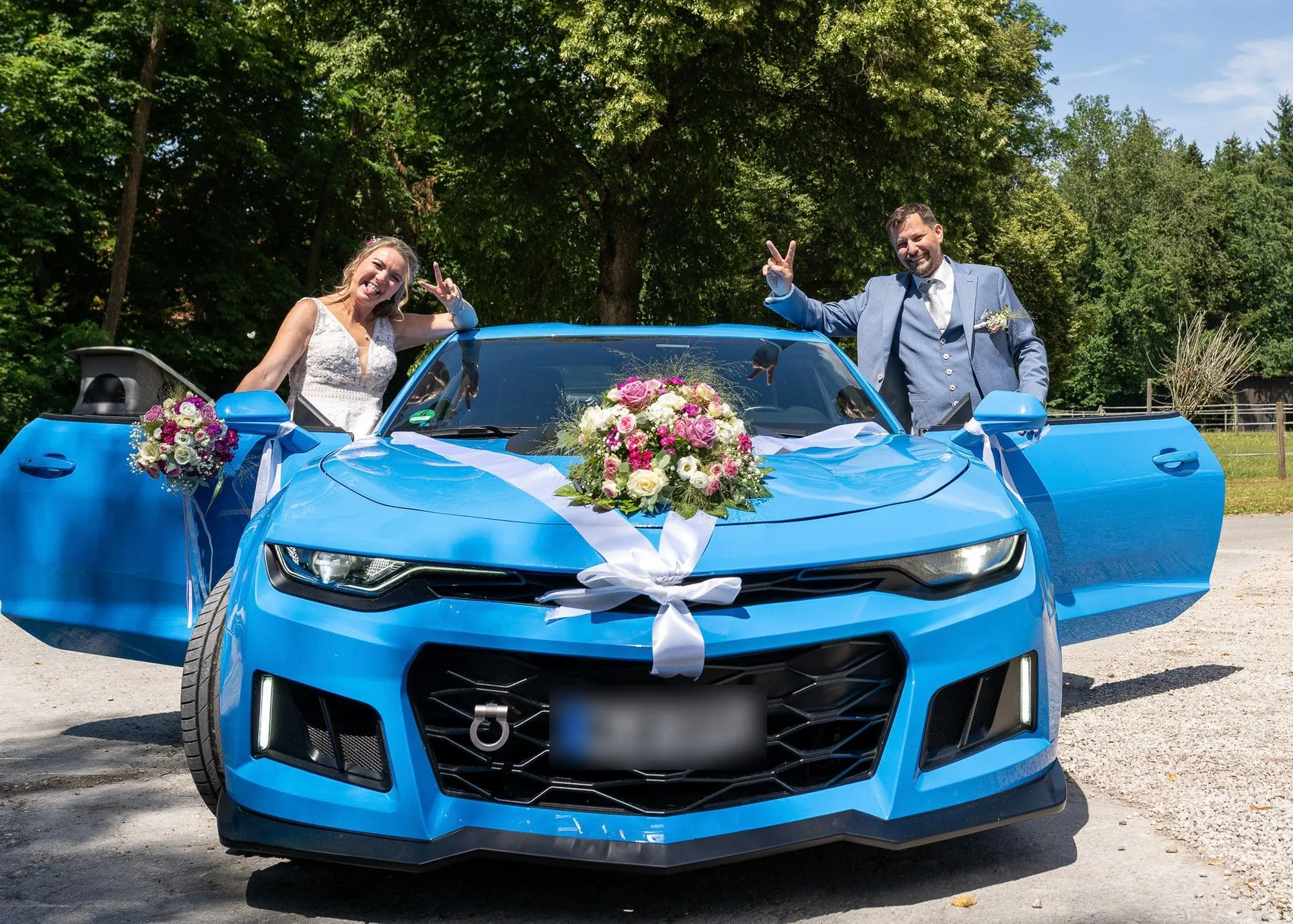 Brautpaar winkt neben dem blauen, mit Blumen geschmückten Hochzeitsauto